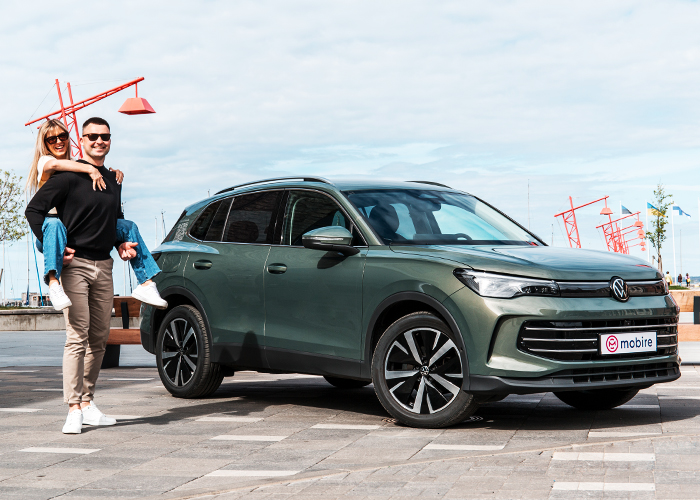 A young couple next to a green SUV with a 'Mobire' logo. The woman is piggybacking on the man, both smiling in a modern outdoor setting