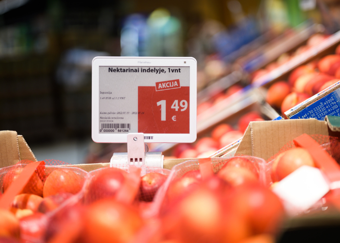 A price tag displaying a special offer for nectarines in a store. The price is shown as 1.49 euros per unit, with the word 'AKCIJA' (meaning 'special offer' in Lithuanian) highlighted in red. Nectarines are visible in red netted bags surrounding the price tag.